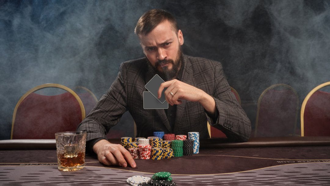 A poker player looks over at his opponent while holding his two hole cards in his hand. There are many stacks of poker chips in front of him and a drink near his right hand.