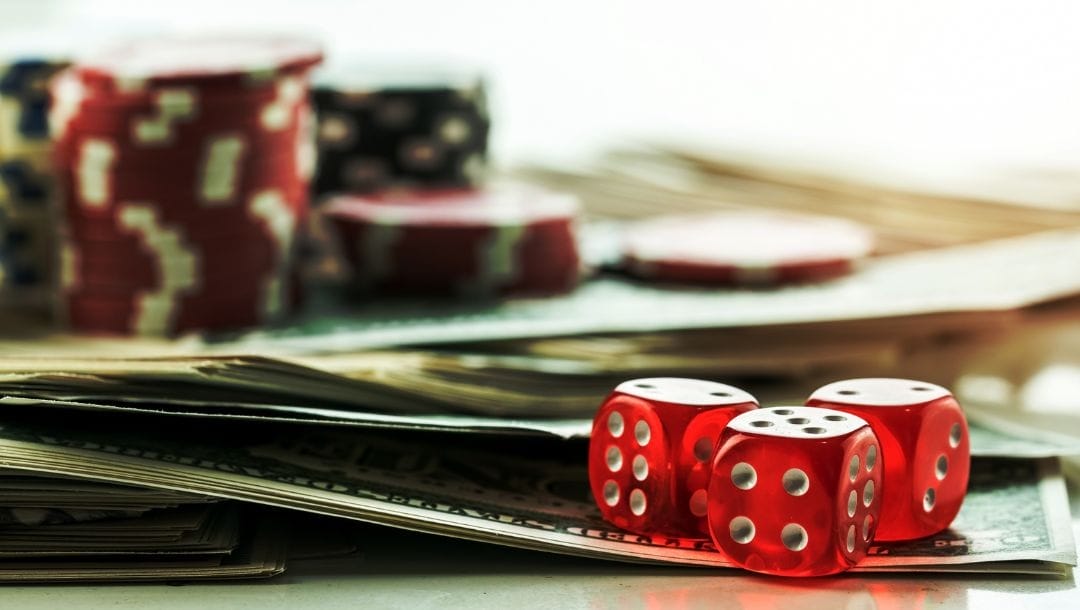 three red six-sided dice on a pile of money bills with poker chips in the blurred background