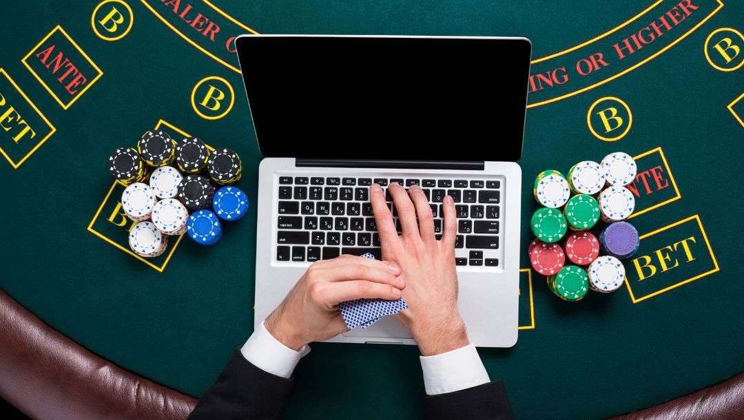 a top view of a man holding two playing cards and typing on a laptop with stacks of poker chips on either side of it on a casino poker table