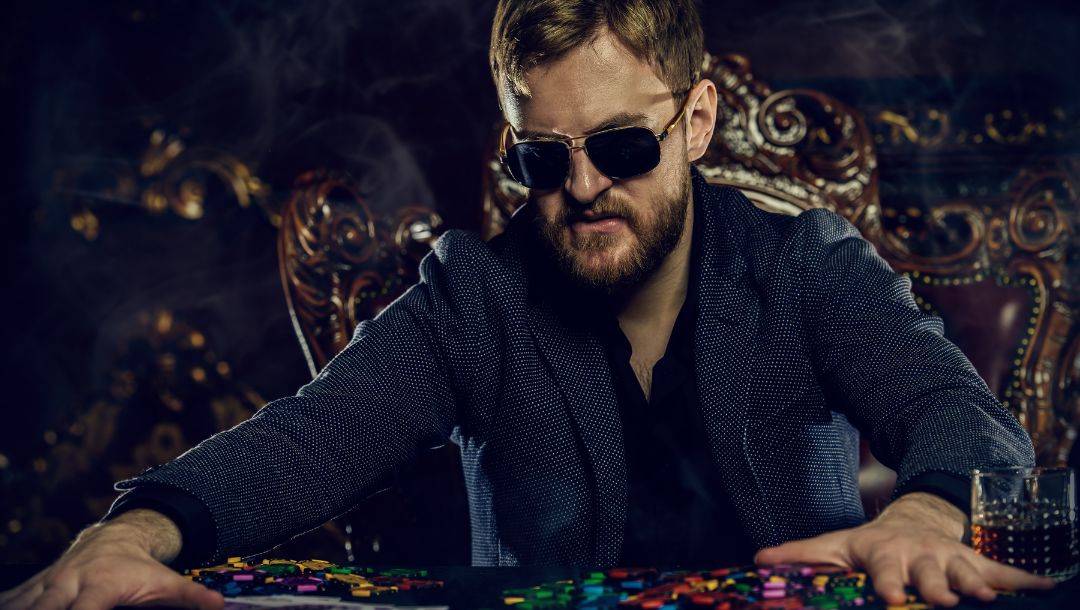 a wealthy man wearing sunglasses is sitting at a poker table and pushing a huge pile of poker chips forward in a casino with smoke in the air