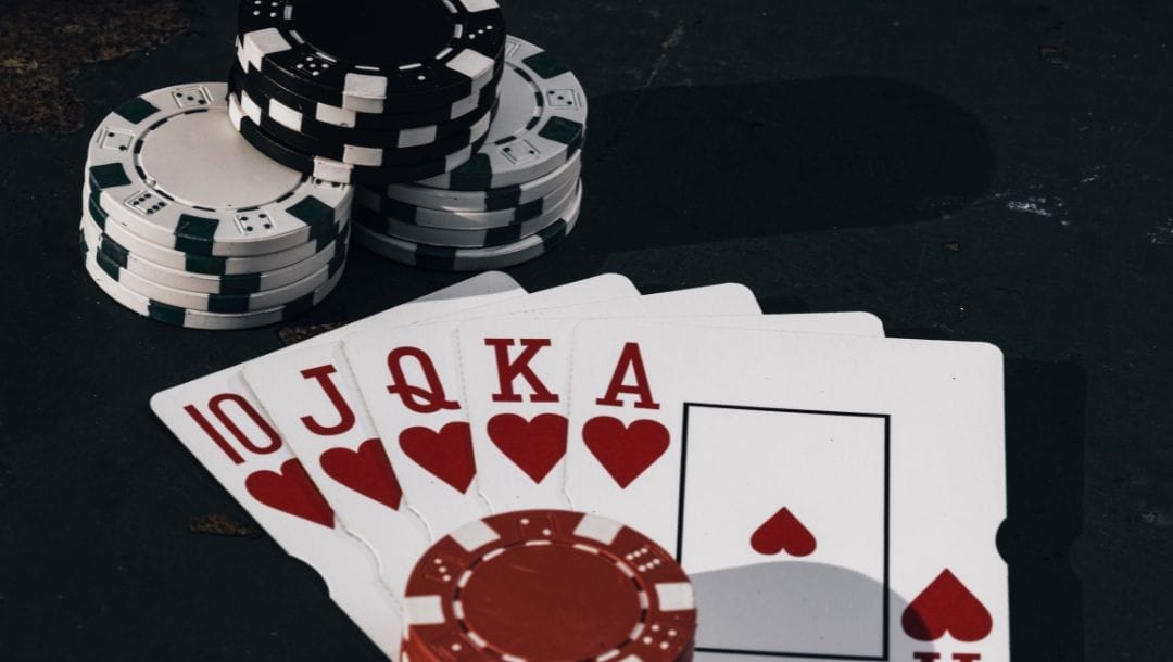 A royal flush of hearts cards fanned out on a black table with red poker chips on top of the cards and black and white poker chips stacked beside the cards.