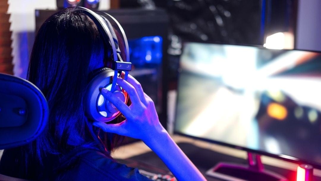 an over-the-shoulder view of a gamer putting on their gaming headset while sitting at a desk and playing video games on a computer