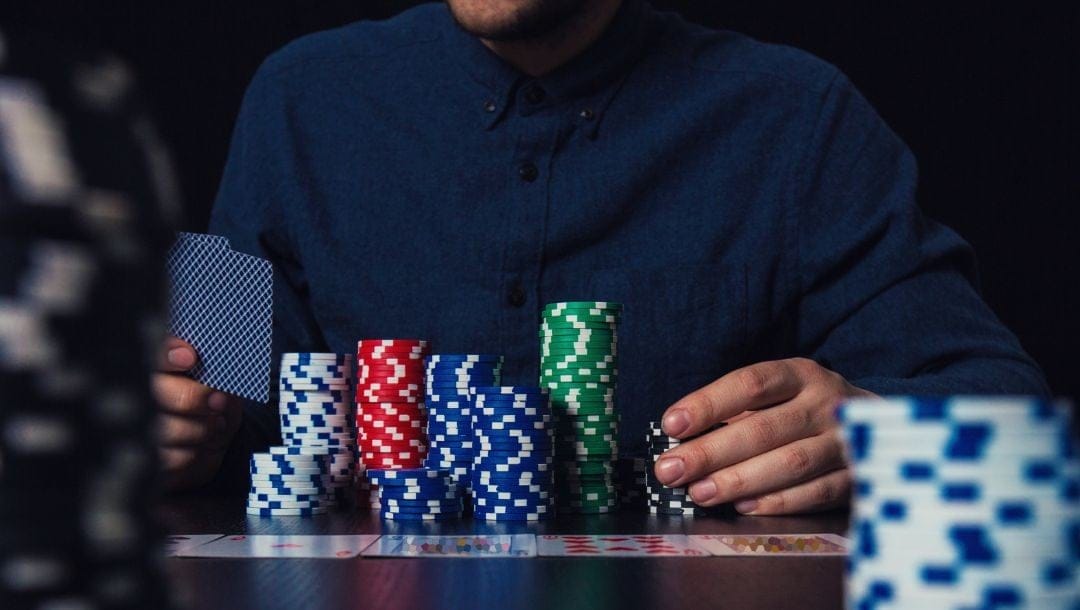 two stacks of poker chips blurred in the foreground, in between them in the background the focus is on a man who is sitting at a table playing poker with poker chips stacked in front of him and two hole cards in his hand, playing cards are laid out face up on the table in front of him
