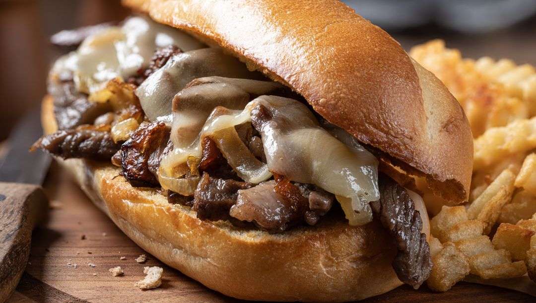 Philly cheesesteak sandwich made with steak, cheese and onions on a toasted hoagie roll with french fries on a wooden board