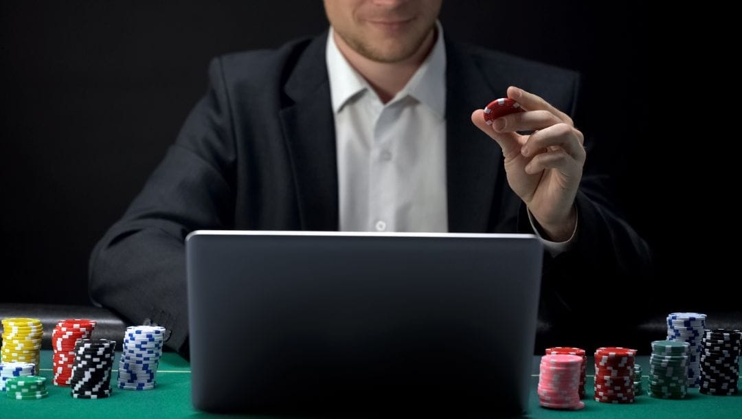 a man holding a red poker chip while sitting in front of a laptop on a green felt poker table with stacks of poker chips on either side of the laptop