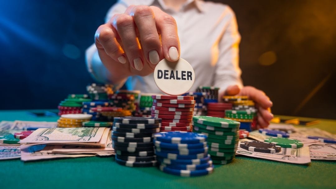 A poker dealer reaching over a stack of poker chips and cash to show the camera their dealer chip.