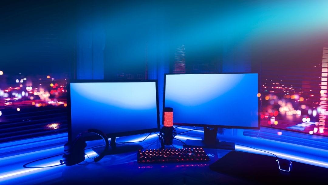 Two PC monitors, a mechanical keyboard, a gaming headset, and a microphone on a table next to a window with a cityscape at night outside, all in ambient neon lighting.