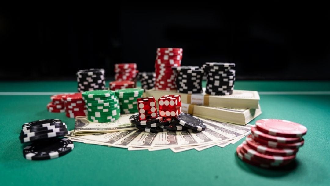 Small stacks of red, black, and green poker chips surround $100 bills layed out on a green table. Two red six-sided dice sit on top of the poker chips in the center of the photograph.