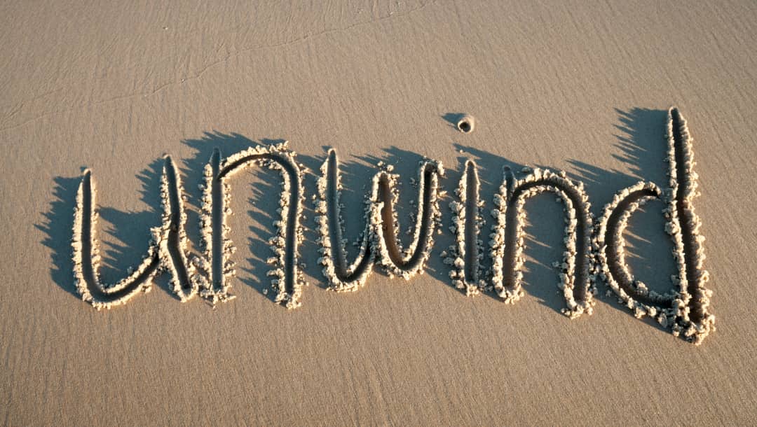 An aerial photograph of the word “Unwind” written in the sand on an empty beach.
