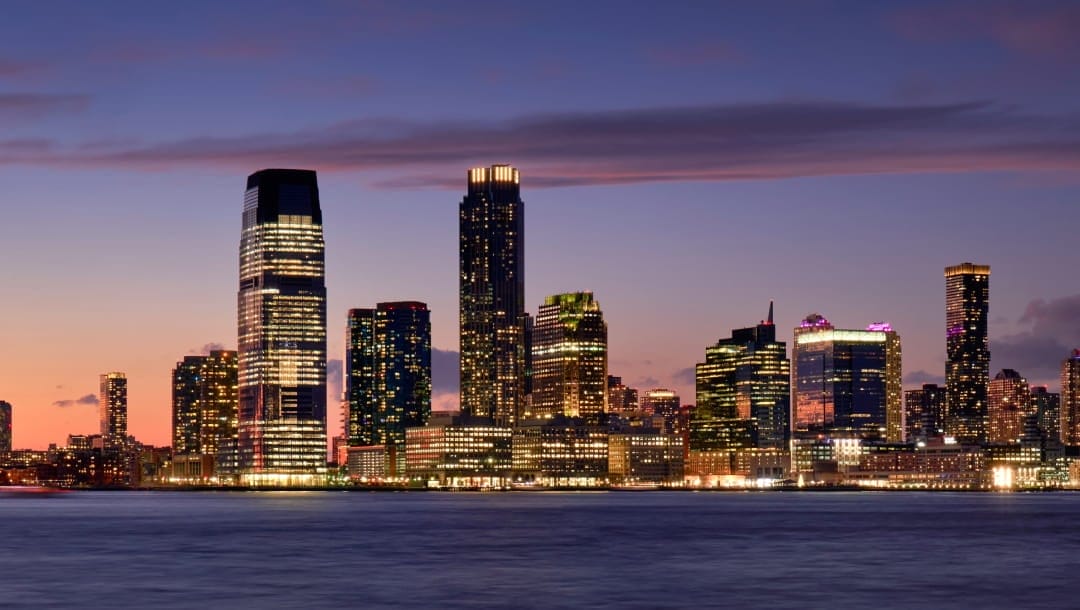 A skyline view of New Jersey City, New Jersey, at night. There’s a bed of water with a purple and orange sky.