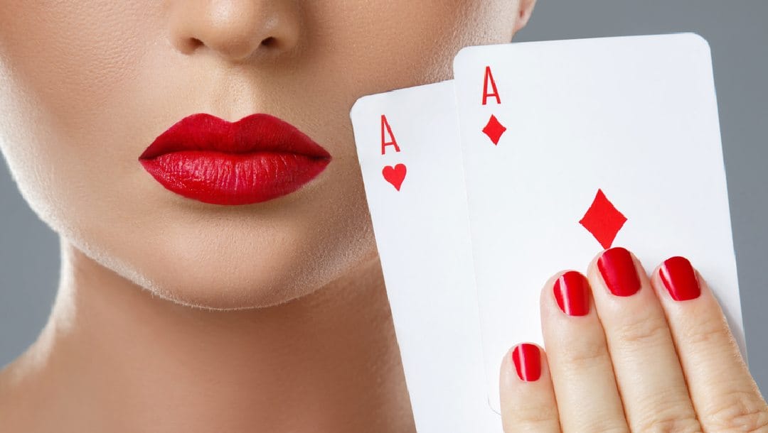 A woman with red lipstick and red nails holding up an ace of hearts and diamonds