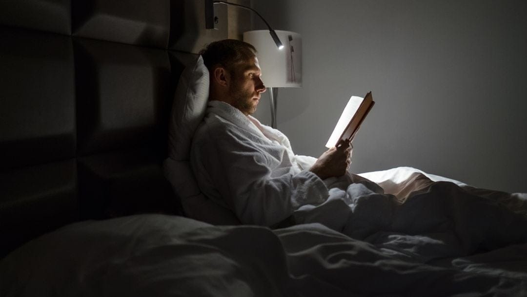Man sitting up in bed, illuminated by a bedside lamp,reading a book.