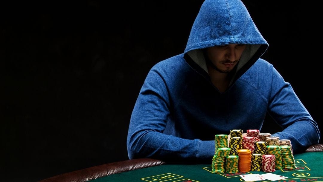 A man in a blue hoodie seated at a poker table, with a stack of cards and casino chips neatly arranged in front of him.