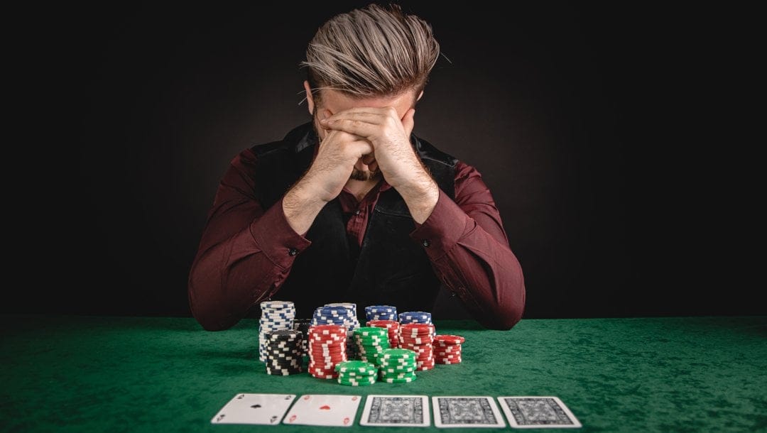 A person holds their head in their hands. A stack of casino chips and five playing cards are on the table in front of them.