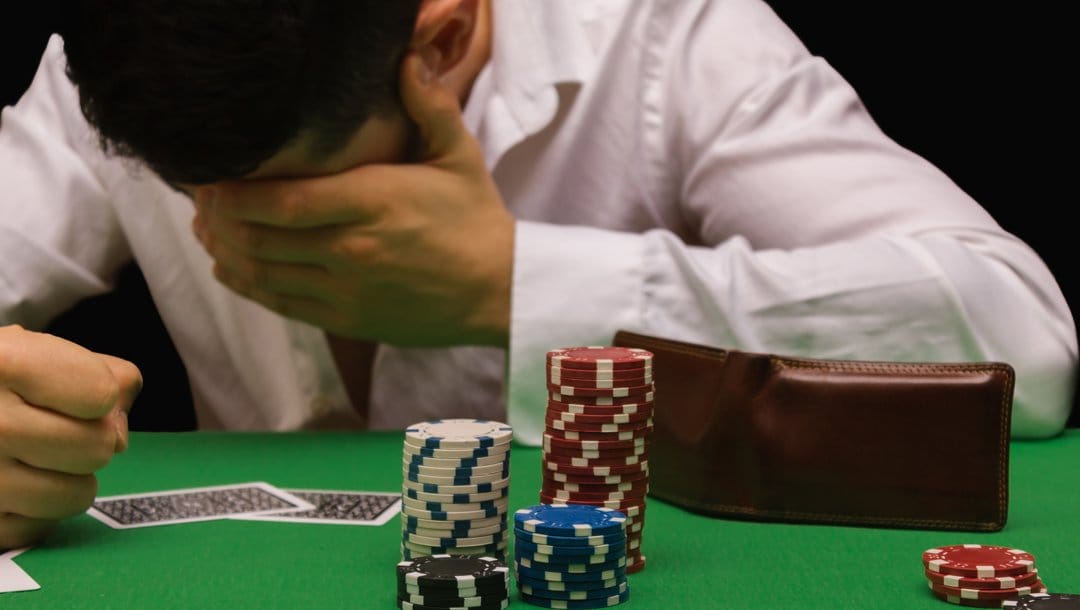 A poker player holds their head in one hand and clenches the other while leaning on a poker table. There are playing cards, stacks of poker chips, and an empty wallet on the table.
