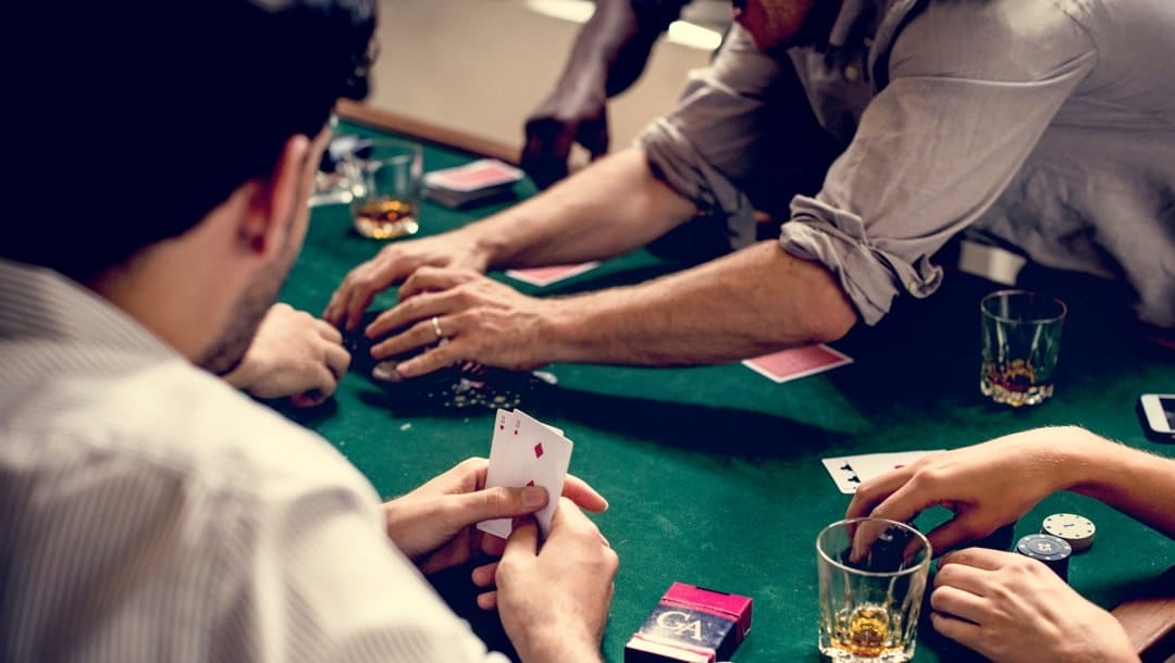 A poker player with their sleeves rolled up reaches over to grab the pot in a poker game. The rest of the players watch.