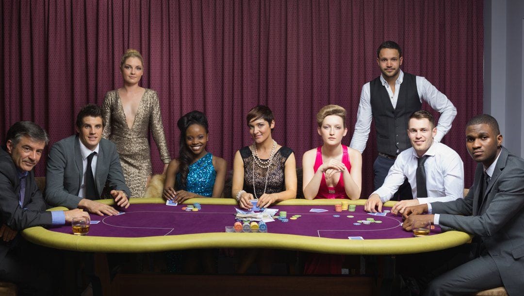 A well-dressed group of people sitting and standing around a poker table.