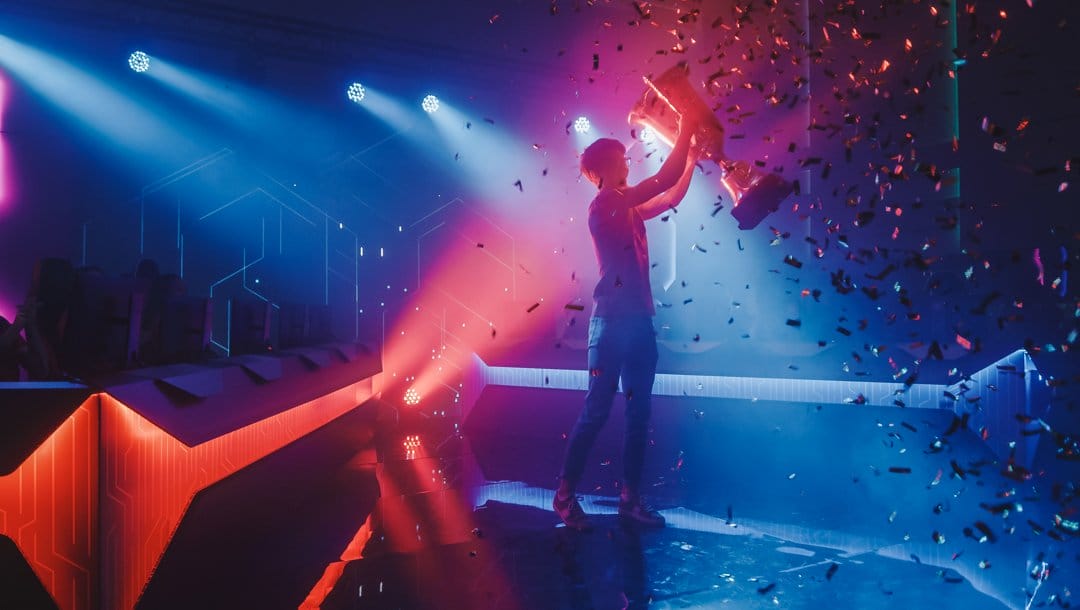 An esports professional holding up a trophy. Ticker tape falls around them while bright red and blue lights shine from behind them.