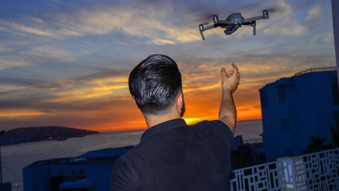 A man standing on his balcony releases his drone at sunset with the ocean in the background.