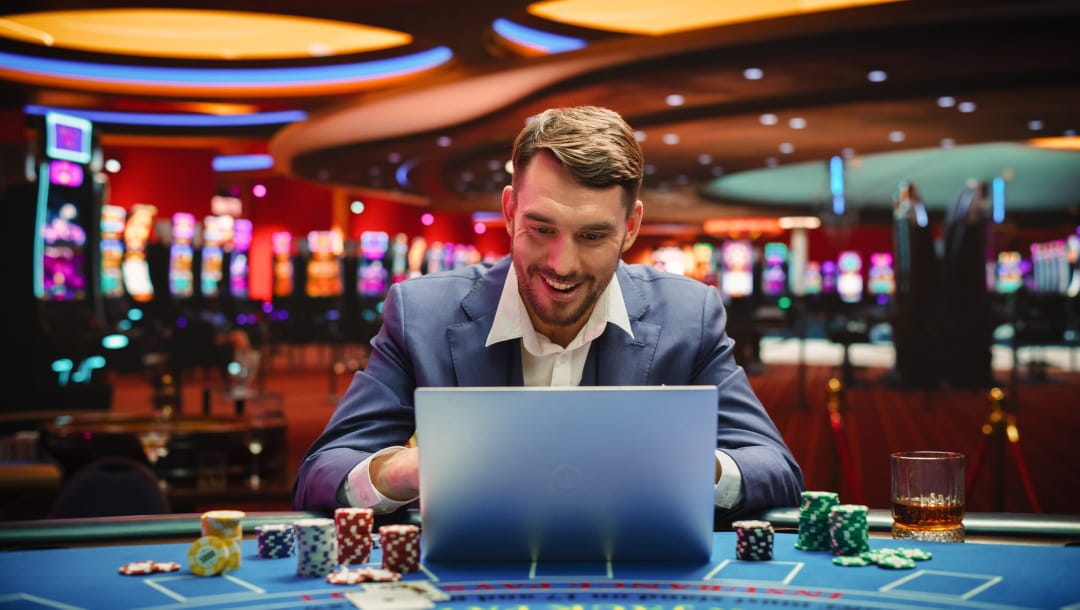A man in a suit smiles while using a laptop at a casino table. There are stacks of casino chips, playing cards, and a drink next to his laptop.
