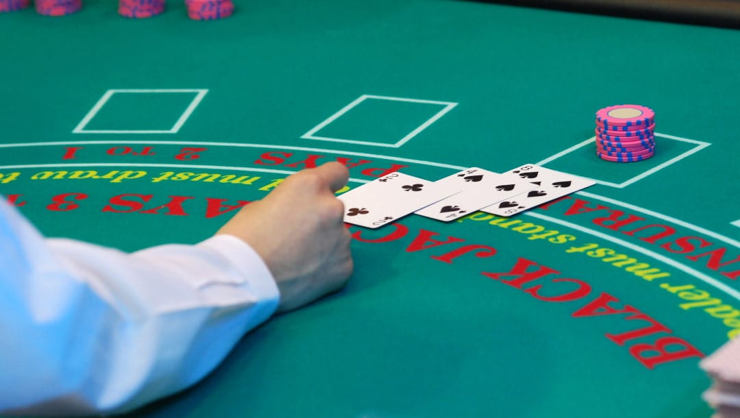 A dealer reaching towards three cards on a green blackjack table. The cards are a two of clubs, a four of spades, and an eight of spades. A stack of pink and blue casino chips sit behind the cards. A few stacks of pink and blue casino chips sit at the edge of the table.