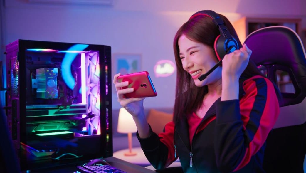 A female gamer sitting in front of her gaming setup, holding her phone and celebrating in ambient purple lighting.