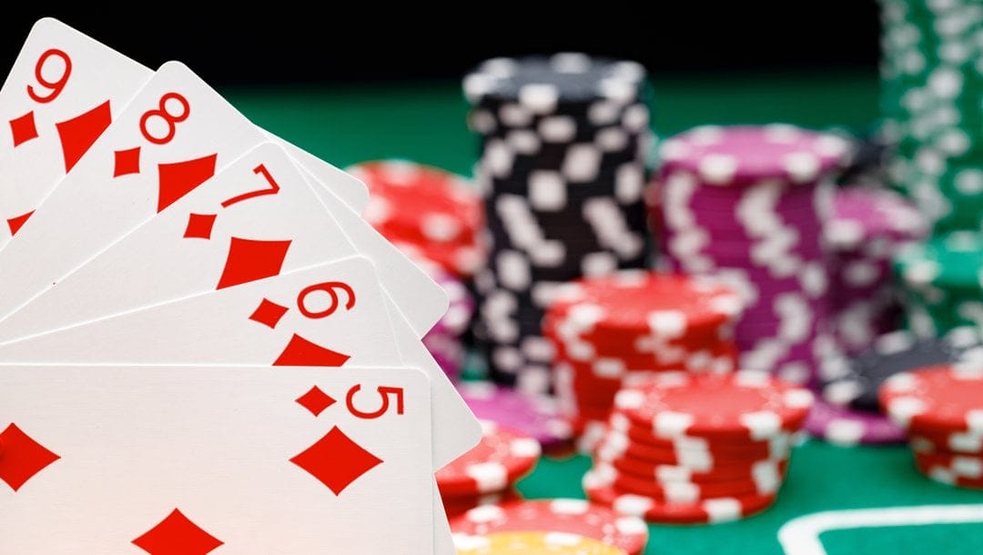 A straight flush held in front of stacks of poker chips on a poker table. The straight flush is the five to nine of diamonds.
