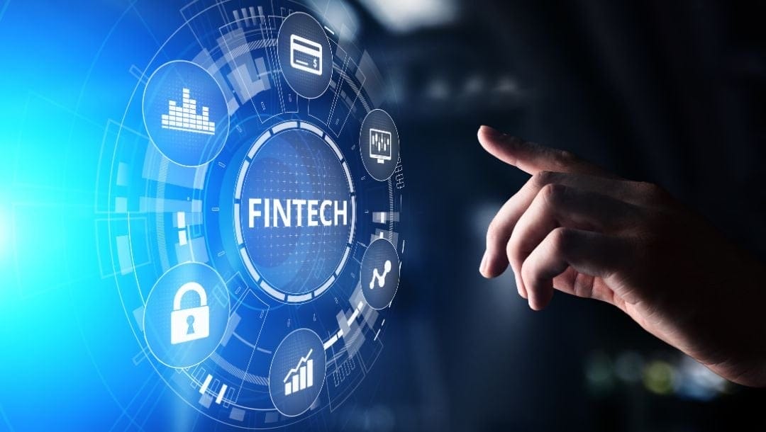 A hand reaches toward a digital, holographic chart with the word “Fintech” in the centre and various financial-technology-related symbols surrounding it.