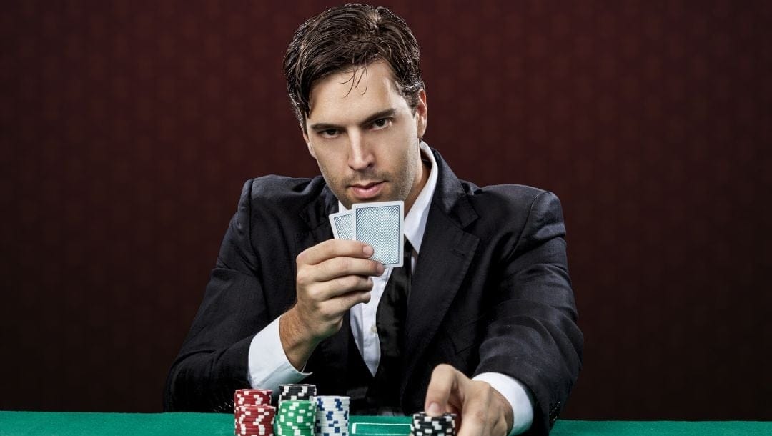 A man wearing a suit, with a very serious expression on his face, seated at a poker table, holding two playing cards in one hand, pushing poker chips forward with the other hand.