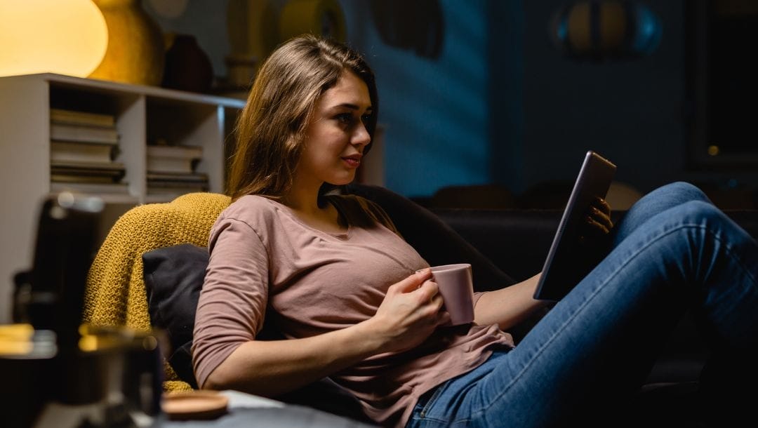 A woman relaxing on her couch drinking a cup of coffee while she plays online casino games on her tablet.