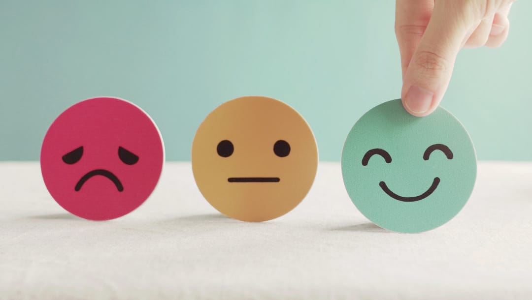 Small emoji cut-outs on top of a white surface. There’s a pink, sad emoji, a yellow, neutral-feeling emoji, and a blue, smiling emoji.