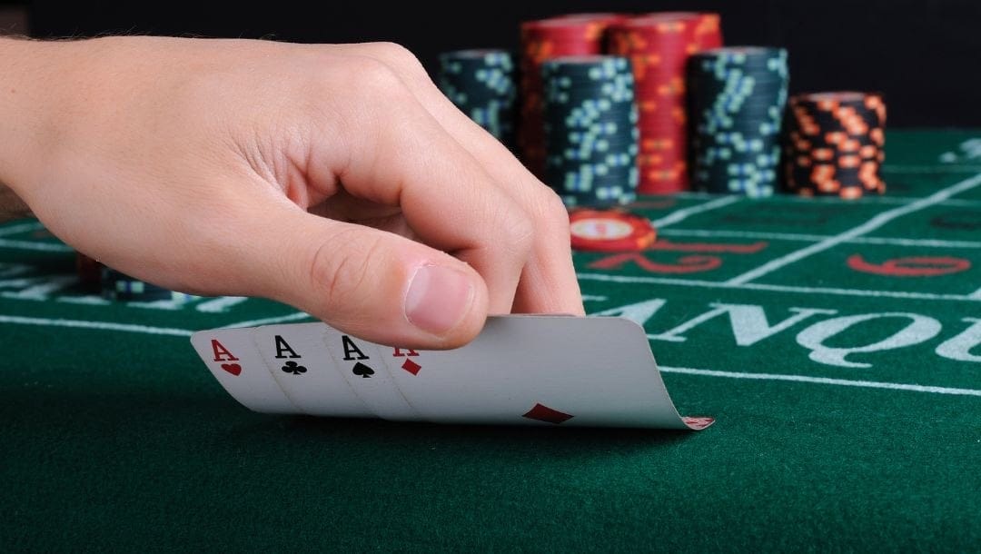 a hand lifting four of a kind ace playing cards off a green felt baccarat table with casino gambling chips on it