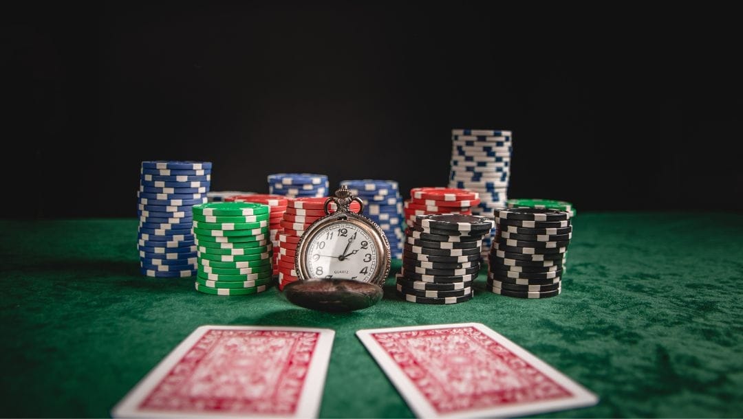 Two playing cards lying face down on a green felt poker table with stacks of poker chips at the rear and a pocket watch in the centre.
