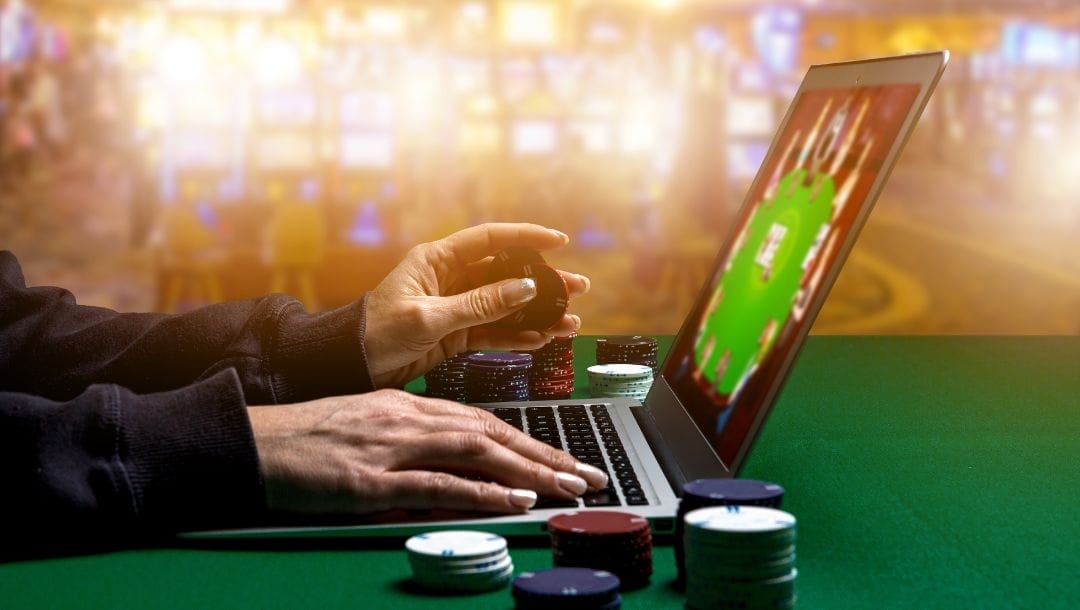 A woman is gambling online on her laptop. She is holding a couple of poker chips and there are a few stacks of poker chips around her on a green felt table. Dramatic lighting in the background.