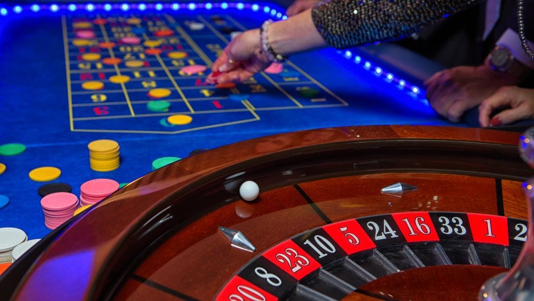 A blue felt roulette table with chips scattered around and a roulette wheel in the forefront.