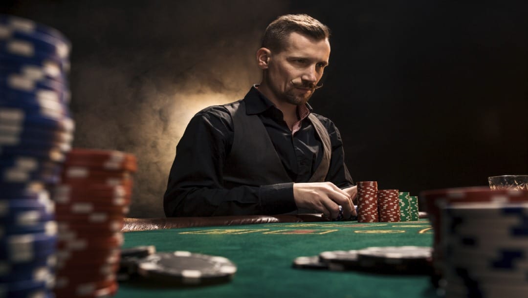 A man sitting at a poker table is surrounded by poker chips, and is thinking about his next move while holding his poker chips.