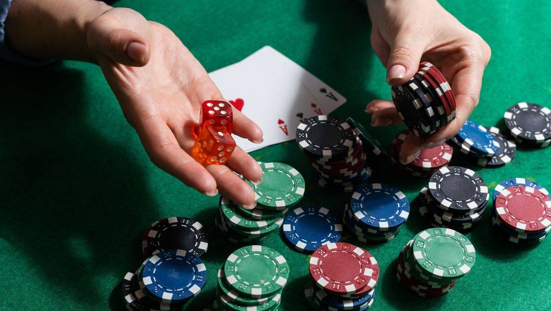 a person holds out a small stack of poker chips and a pair of red dice over a green felt poker table with poker chips and playing cards on it