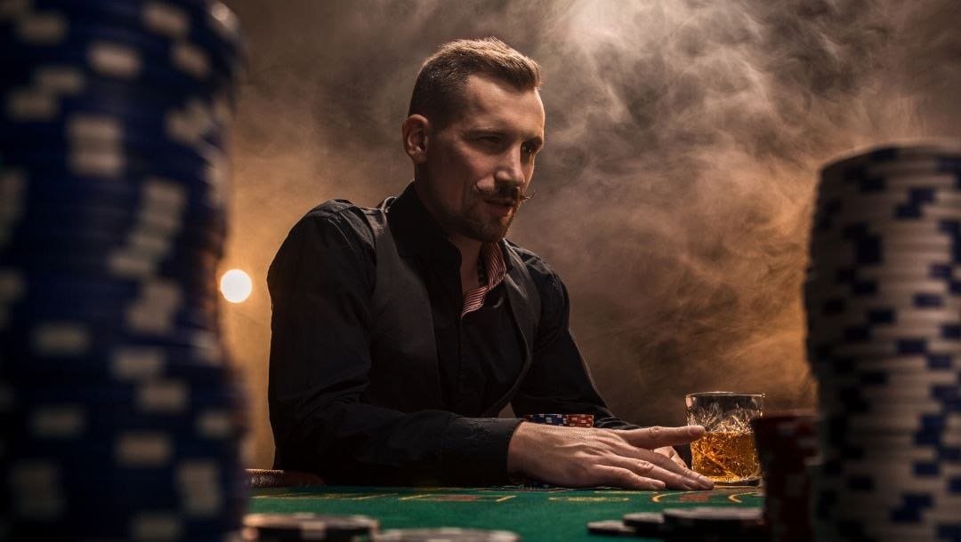 a man is sitting at a poker table with poker chips and a drink in front of him and smoke in the background, the shot is taken through two towers of poker chips at a low angle