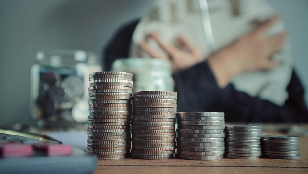 Stacks of coins arranged in ascending order on a wooden table, with jars filled with more coins in the background. A person, partially out of focus, is seen hugging a pillow or soft object, suggesting a theme of saving or financial planning.