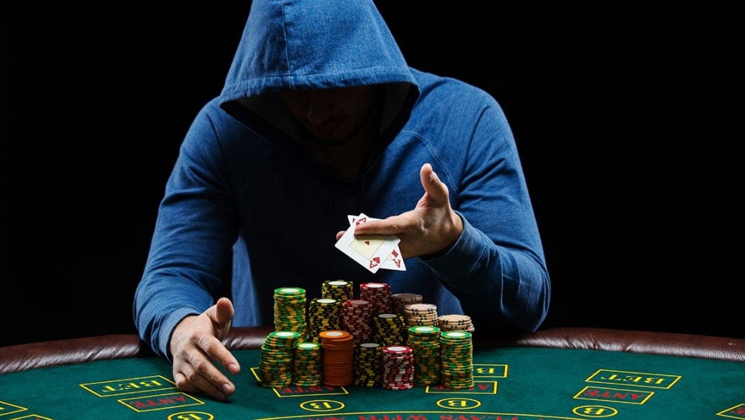 A blackjack player wearing a blue hoodie sits at a casino table under dramatic lighting, partially hiding their face in shadow, with stacks of casino chips in front of them.