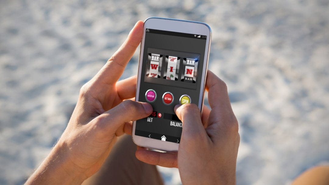 Person playing a slot machine game on a smartphone while sitting on the beach. The screen shows spinning reels and a spin button.