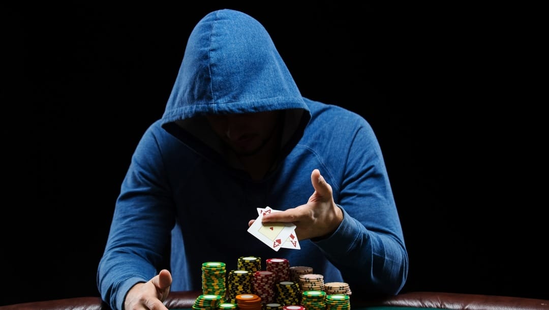 A hooded man hands out two aces, sitting behind many stacks of poker chips