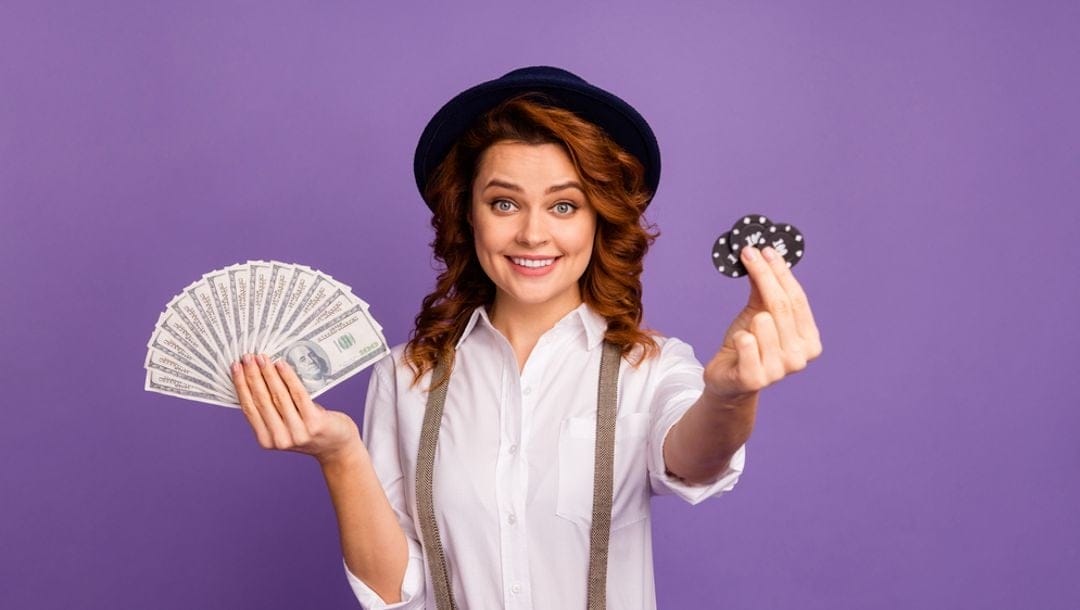 A woman wearing suspenders and a hat holding up casino chips and dollar bills.