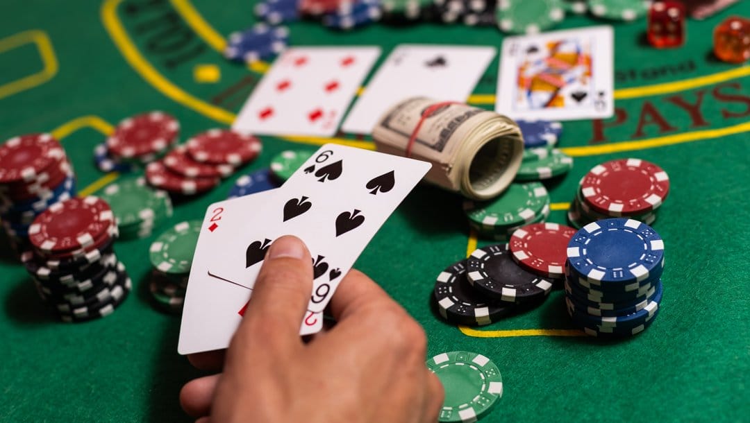 A person holding two playing cards over a casino table. There are playing cards, casino chips, and a roll of cash on the table.