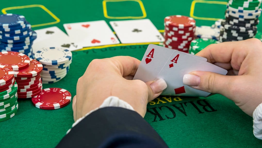 A poker player checks their hole cards. They have a pair of aces. They also have multiple stacks of poker chips on either side of their hands.