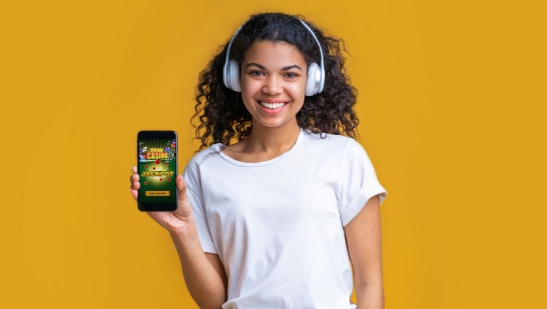 A young, smiling woman listening to music through headphones with her cellphone showing an online casino.