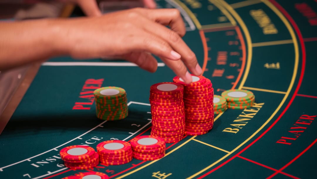 A hand reaching for red casino chips stacked on a green baccarat table, with the "PLAYER" and "BANKER" sections labeled in red and yellow. Additional green and orange chips are scattered around the betting areas.
