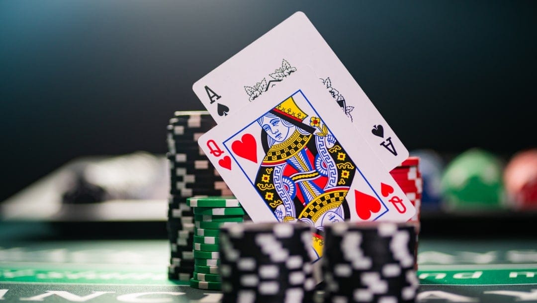 A queen and ace playing card propped up in between casino playing chips on a playing table with a dark background