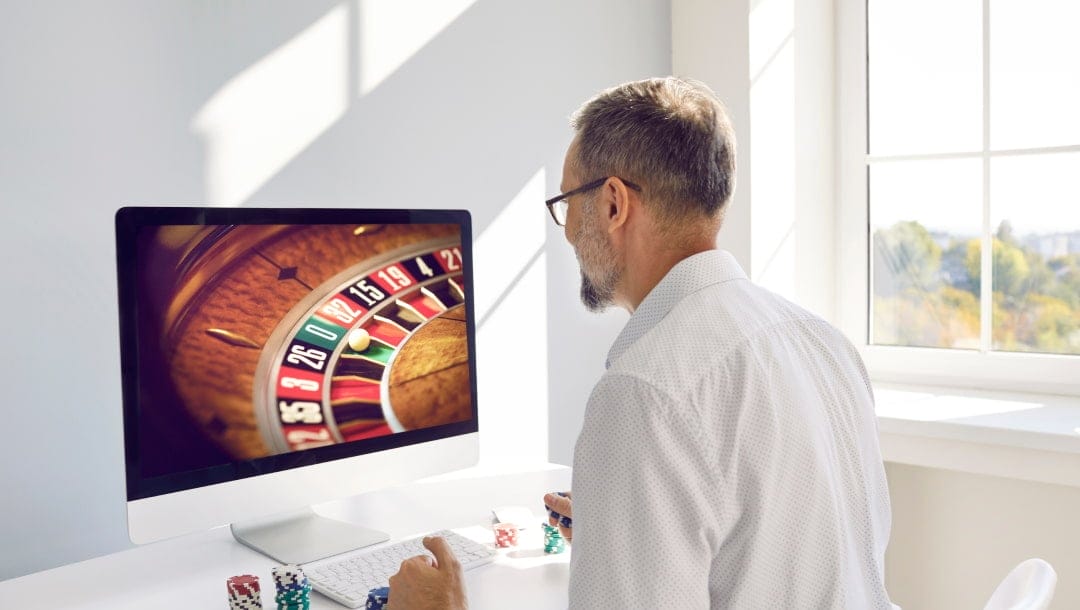 An older man wearing glasses and playing online roulette on his computer in a bright white room. There are stacks of casino chips on either side of his keyboard.