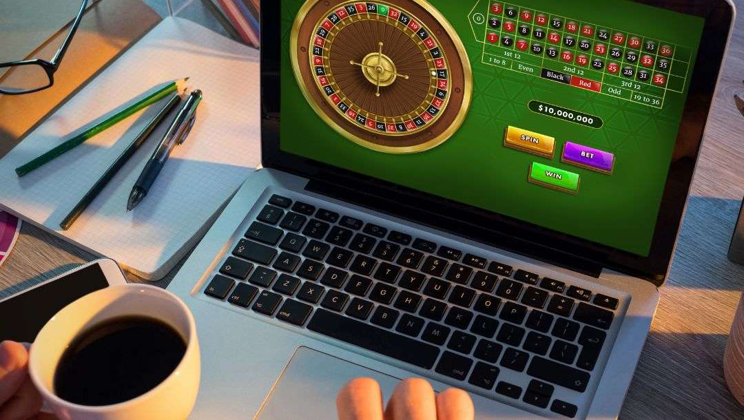 A person sitting at a desk, holding a cup of coffee, playing online roulette on a laptop.